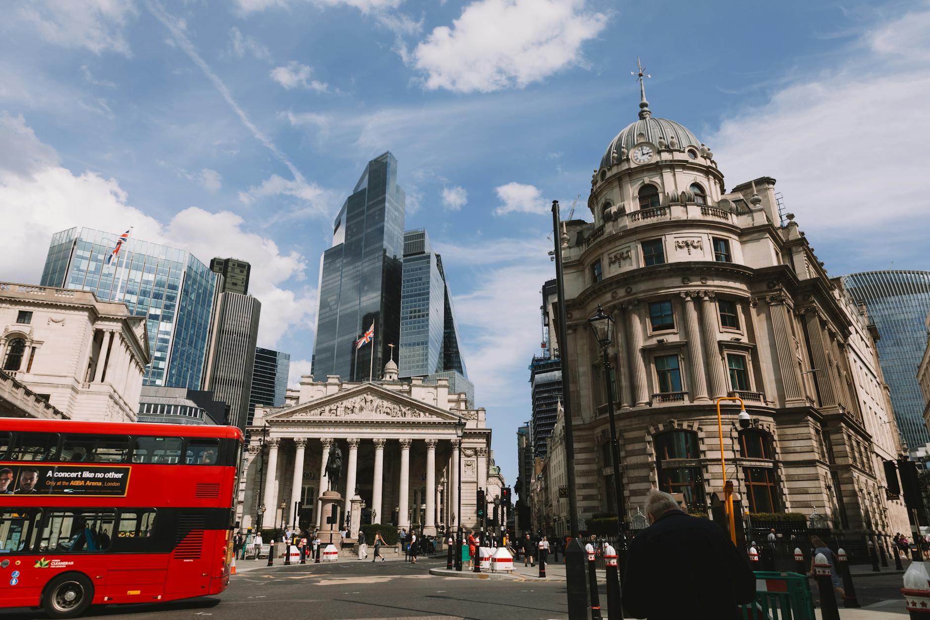 Red London double-decker bus passing the Royal Exchange and Bank of England with City skyscrapers under a blue sky.