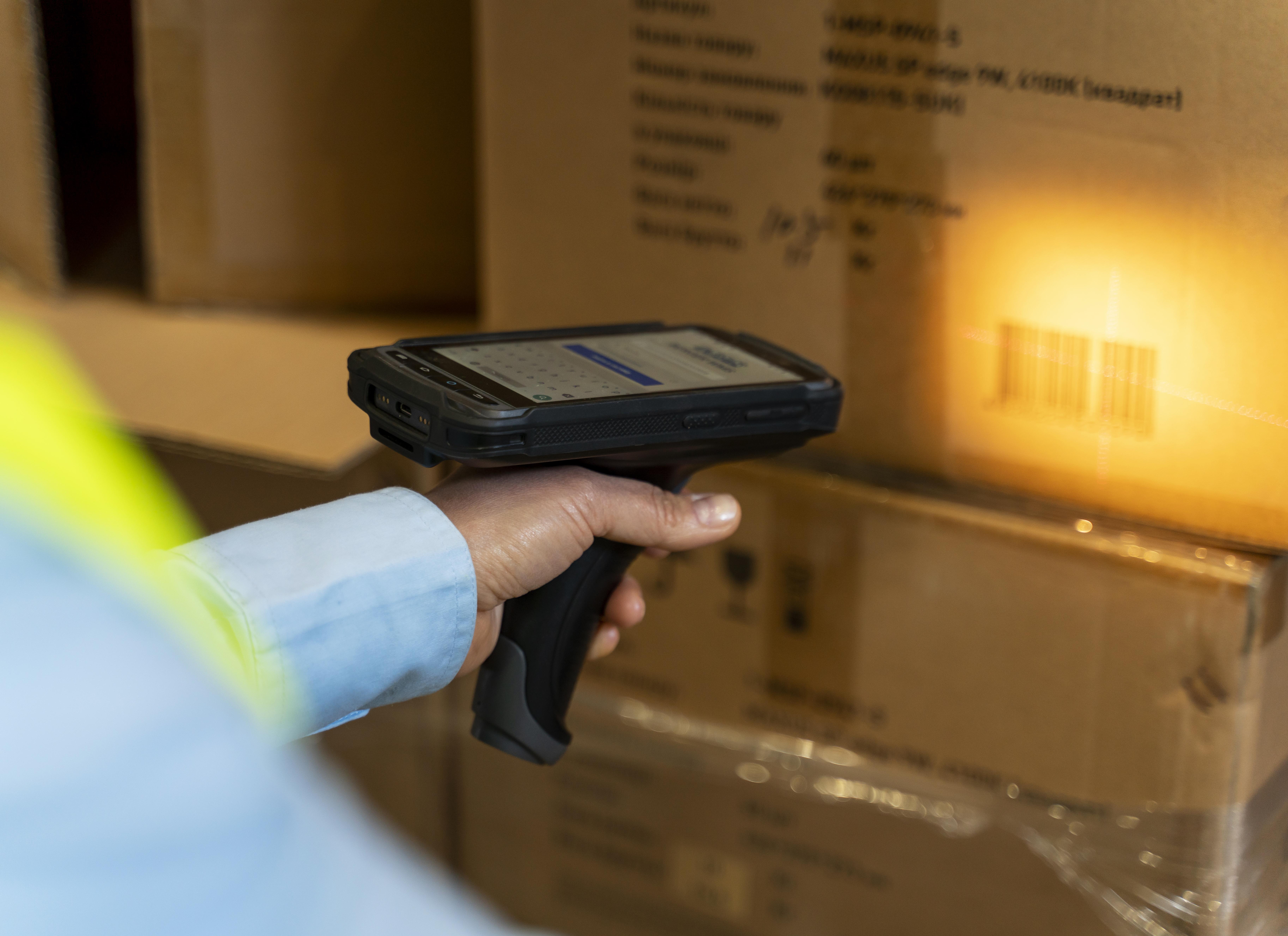 A person's hand holds a barcode scanner pointing at a cardboard box with a glowing barcode. The scene is in a warehouse, suggesting inventory work.