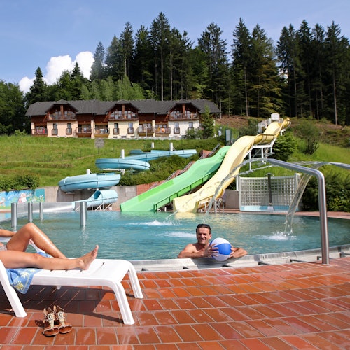 Човек се отпуска на шезлонг близо до басейн с пързалки. Мъж във водата държи синя топка. В гората и сградите на заден план.