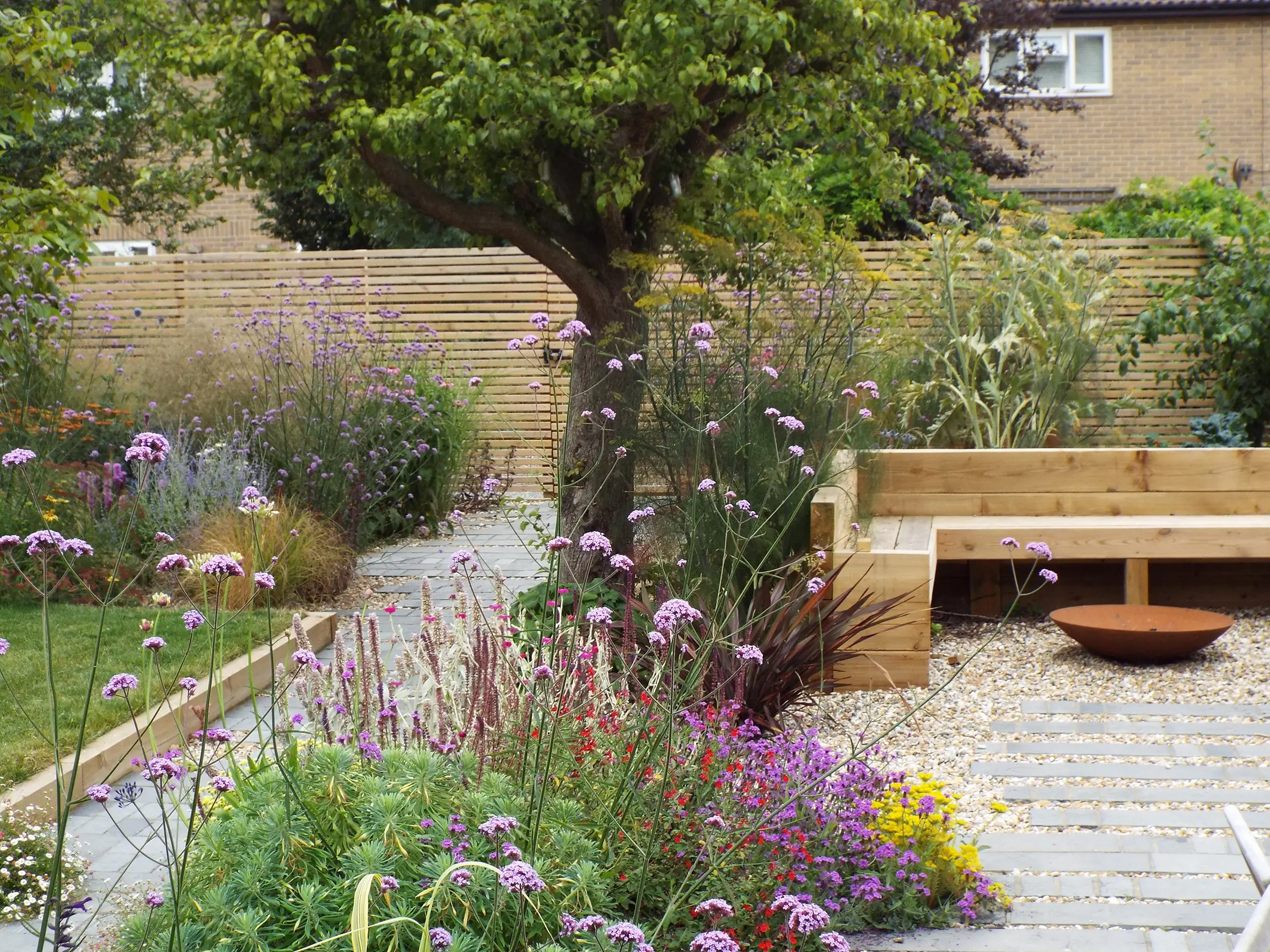 A lush garden scene with a tree, colorful flowers, and a wooden seating area along a stone path.