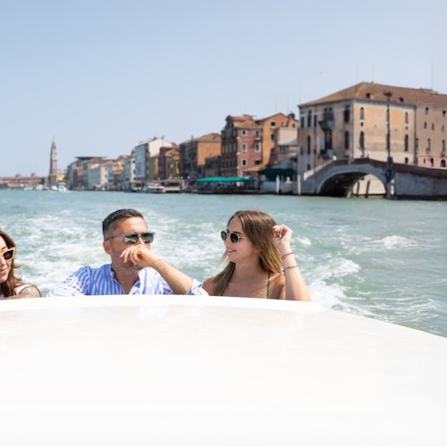 Three people on a boat in a canal with historic buildings and a distant bell tower in the background on a sunny day.