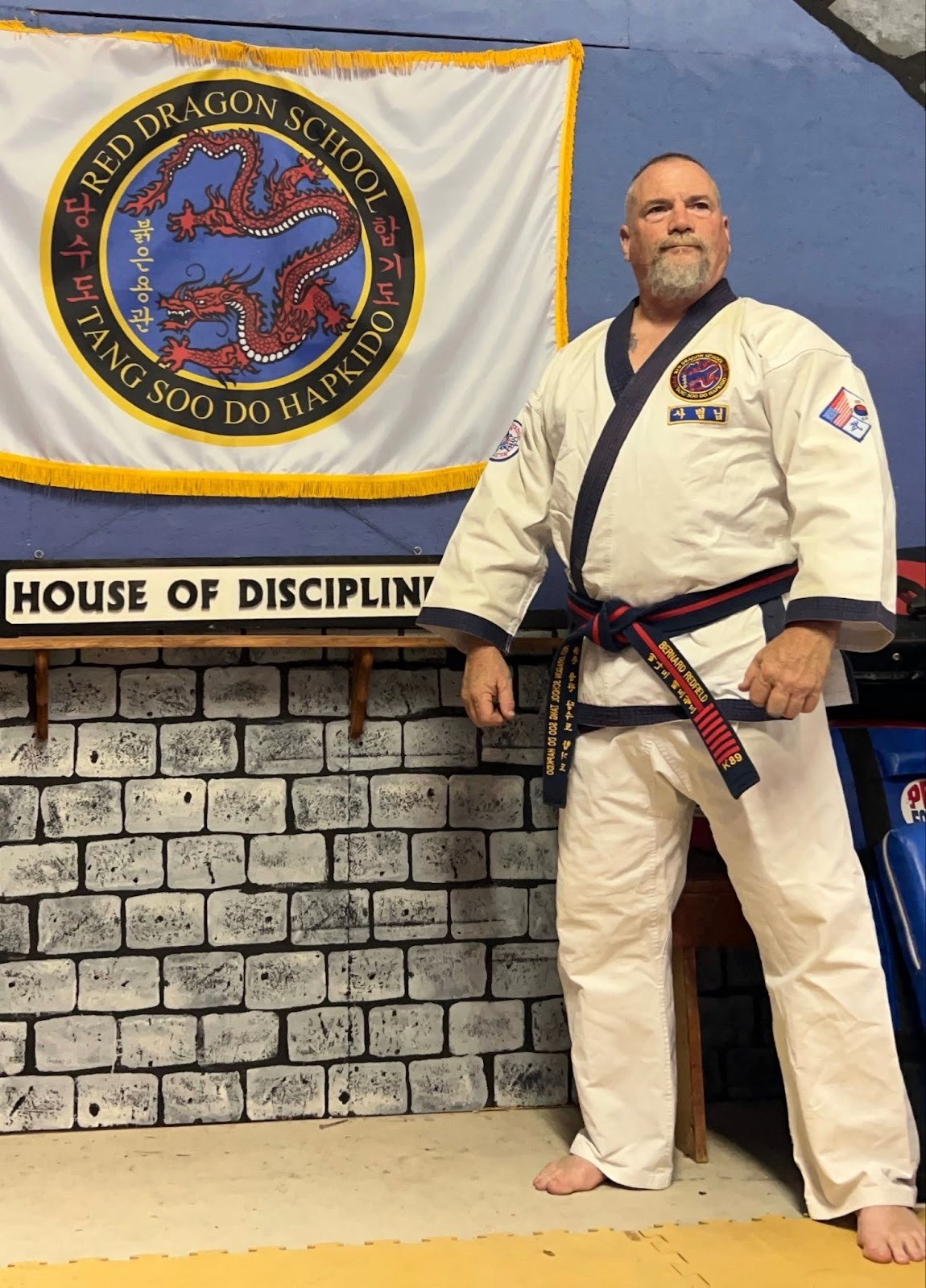A man in a karate uniform stands proudly in front of a "House of Discipline" banner at Redfield Martial Arts school.