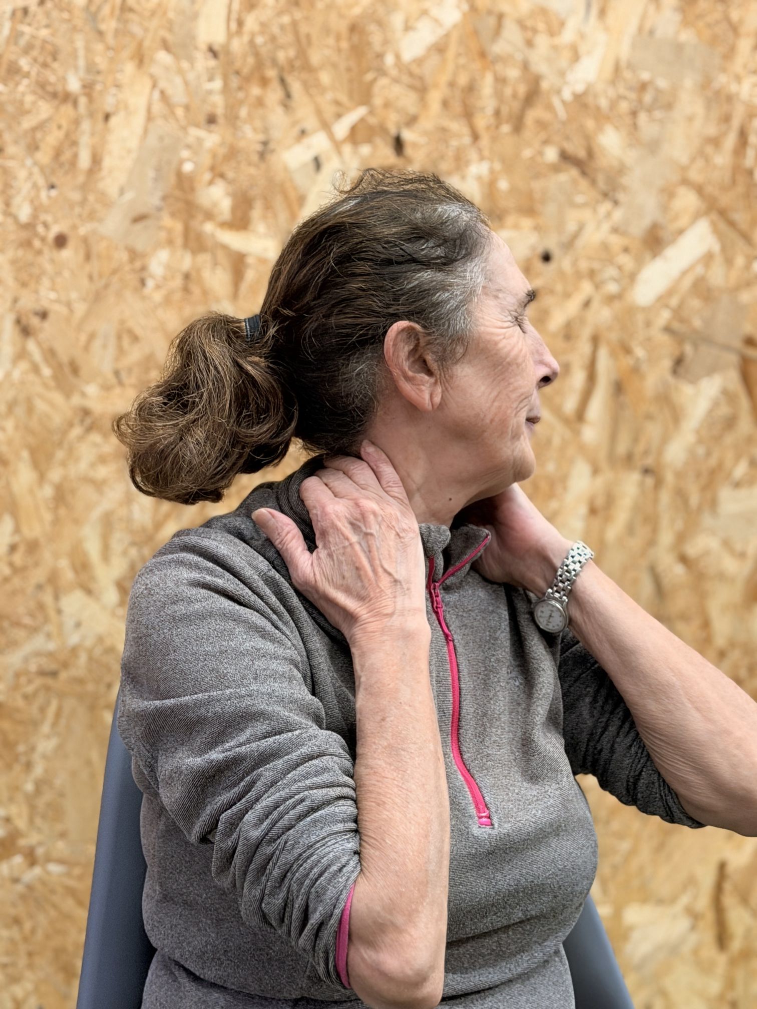 Senior woman in gray activewear top with pink zipper, massaging neck.