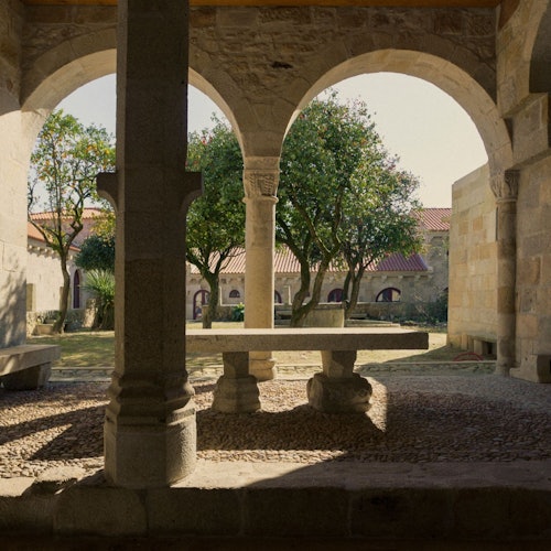 Arcos de piedra enmarcando un patio con árboles, un banco de piedra y una mesa de madera. La luz del sol proyecta sombras en el suelo de piedras.