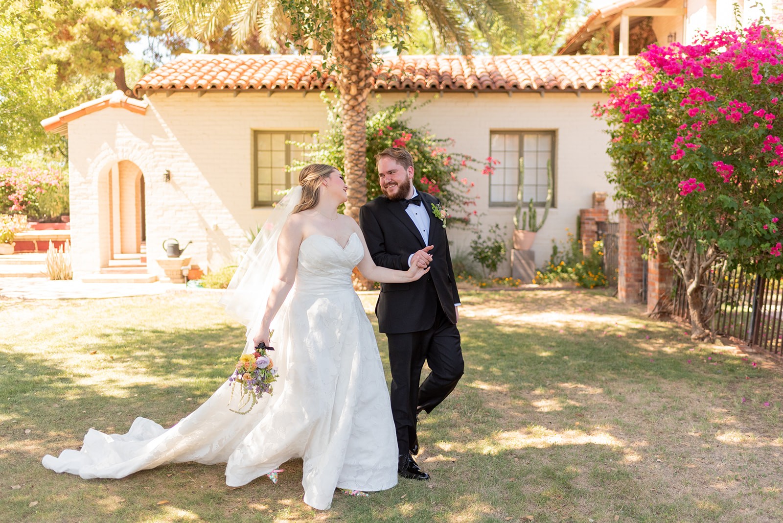 Tess and Alex smile and laugh with a house and blooming flowers behind them just prior to their wedding.