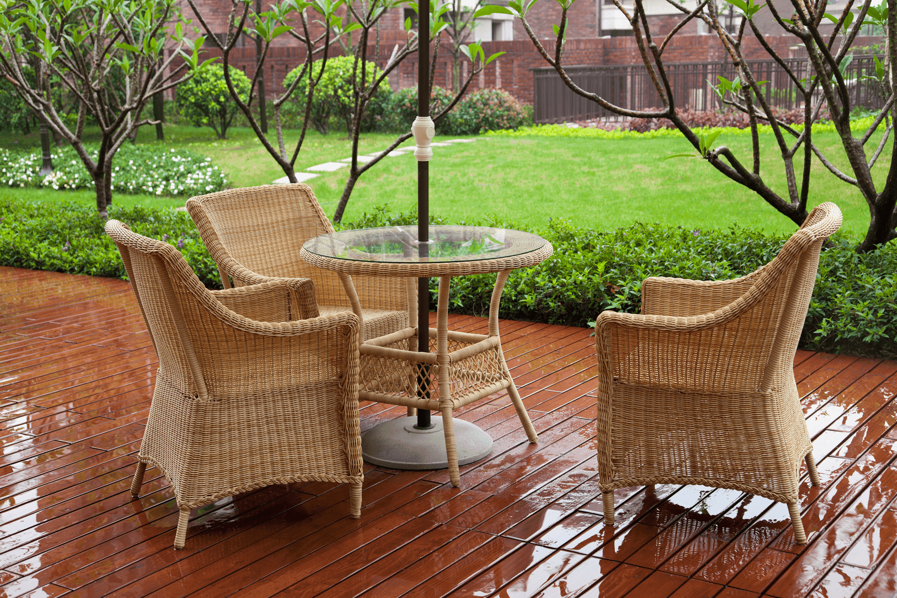 A cozy wooden patio with a glass-top table and four wicker chairs, set on a polished surface glistening from recent rain. The patio is surrounded by neatly trimmed hedges and vibrant greenery in a residential setting.