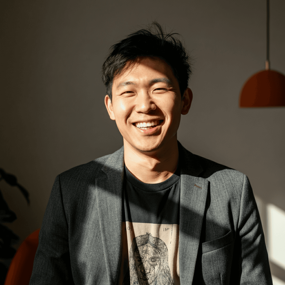 Smiling Asian man in a gray blazer and black t-shirt with artistic design, warmly lit by natural light.