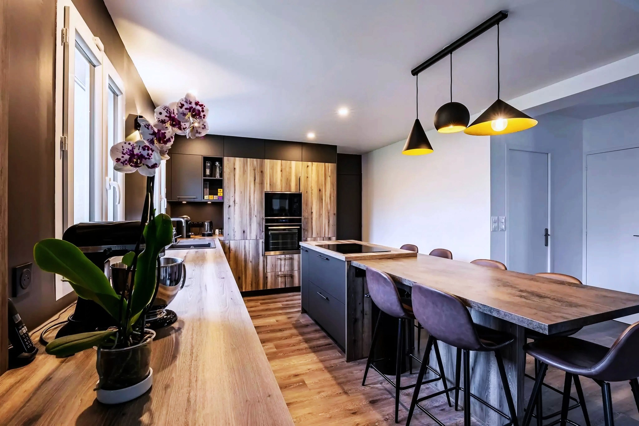 Cuisine moderne avec des comptoirs en bois, un îlot avec des tabourets de bar, des lampes suspendues, des fours encastrés, des armoires sombres et une orchidée en pot sur le comptoir. L'espace est lumineux et contemporain.