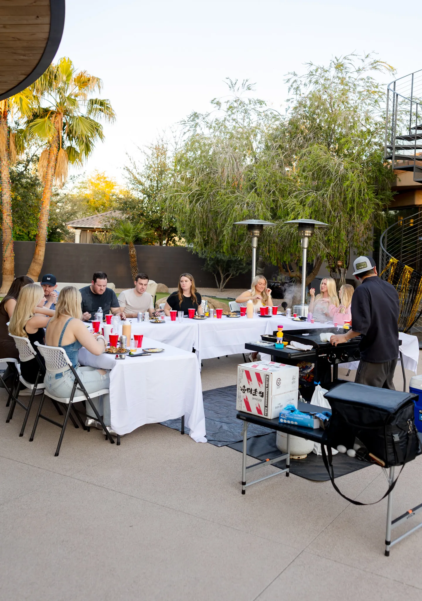 A bright, modern dining room featuring a long light wood table set with several matching wooden chairs and white cushions, illuminated by natural light streaming through large windows overlooking a lush green and sunlit yard.