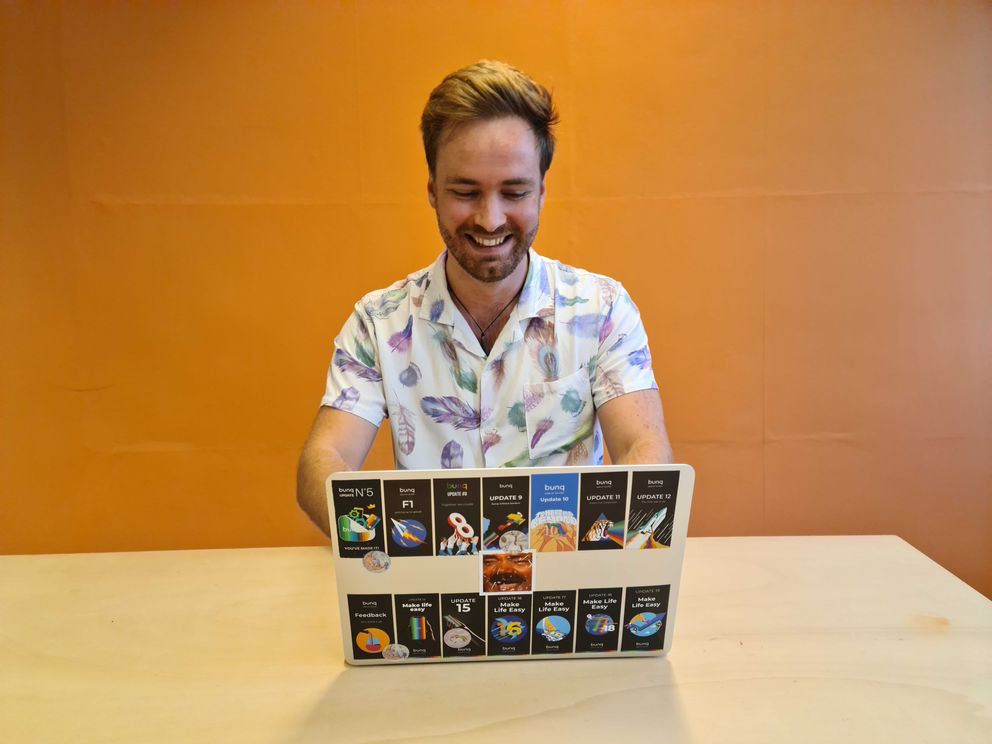 Iemand met kort haar en een bloemenshirt lacht terwijl hij op een laptop werkt aan een tafel, met een oranje muur erachter.