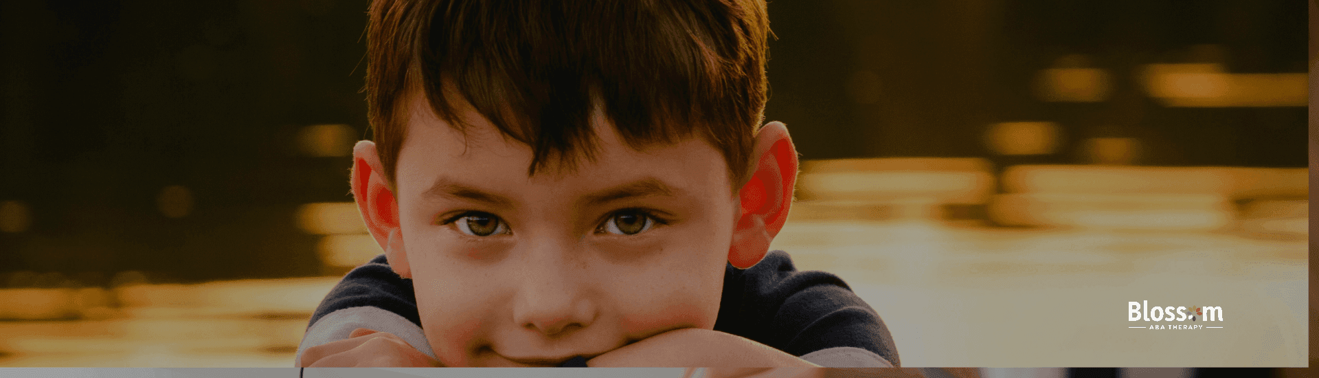 Smiling young boy resting his arms on a railing by a lake at sunset, warm golden light reflecting off the water behind him.