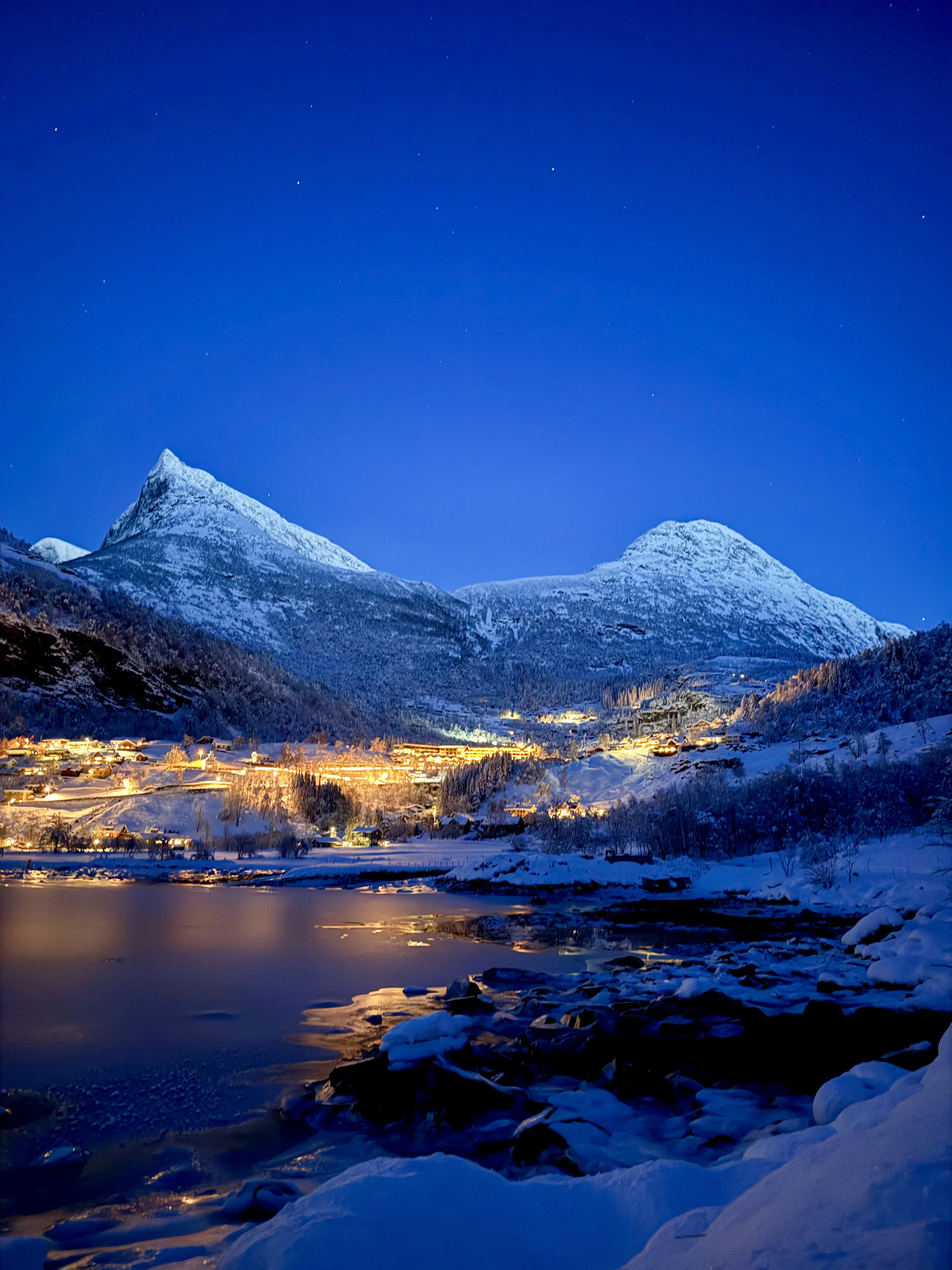 Snow-capped mountains illuminated by twilight, reflecting in calm waters below, creating a serene winter scene.