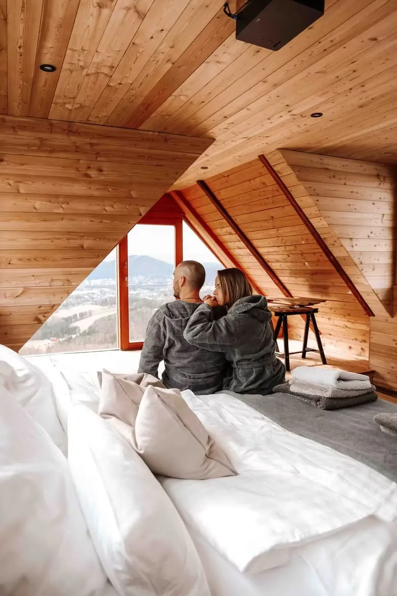 Paar in Bademänteln sitzt auf dem Bett und blickt durch das Giebelfenster ins Tal