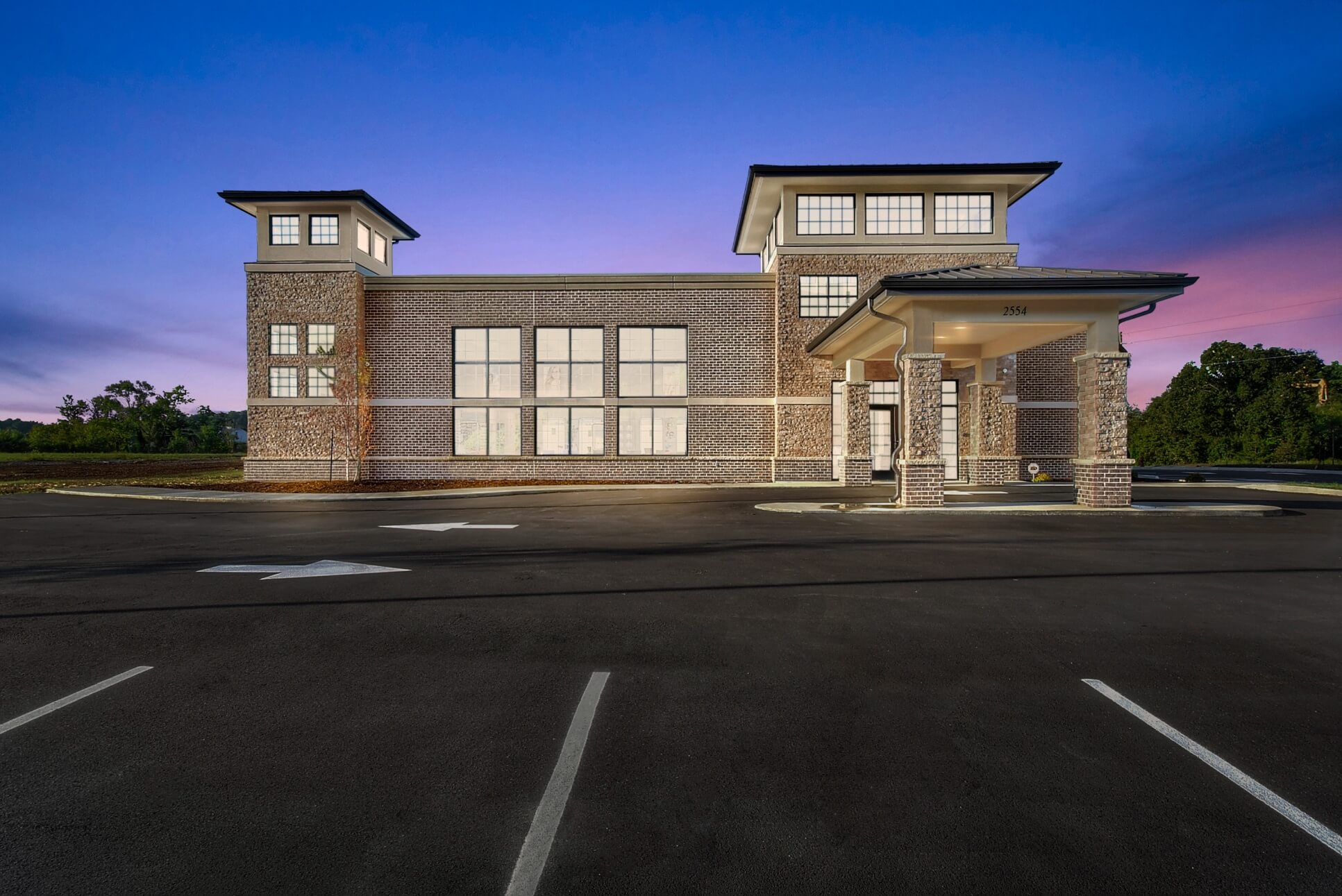 A modern building at dusk, featuring large windows and a welcoming entrance, with an empty parking lot in front.
