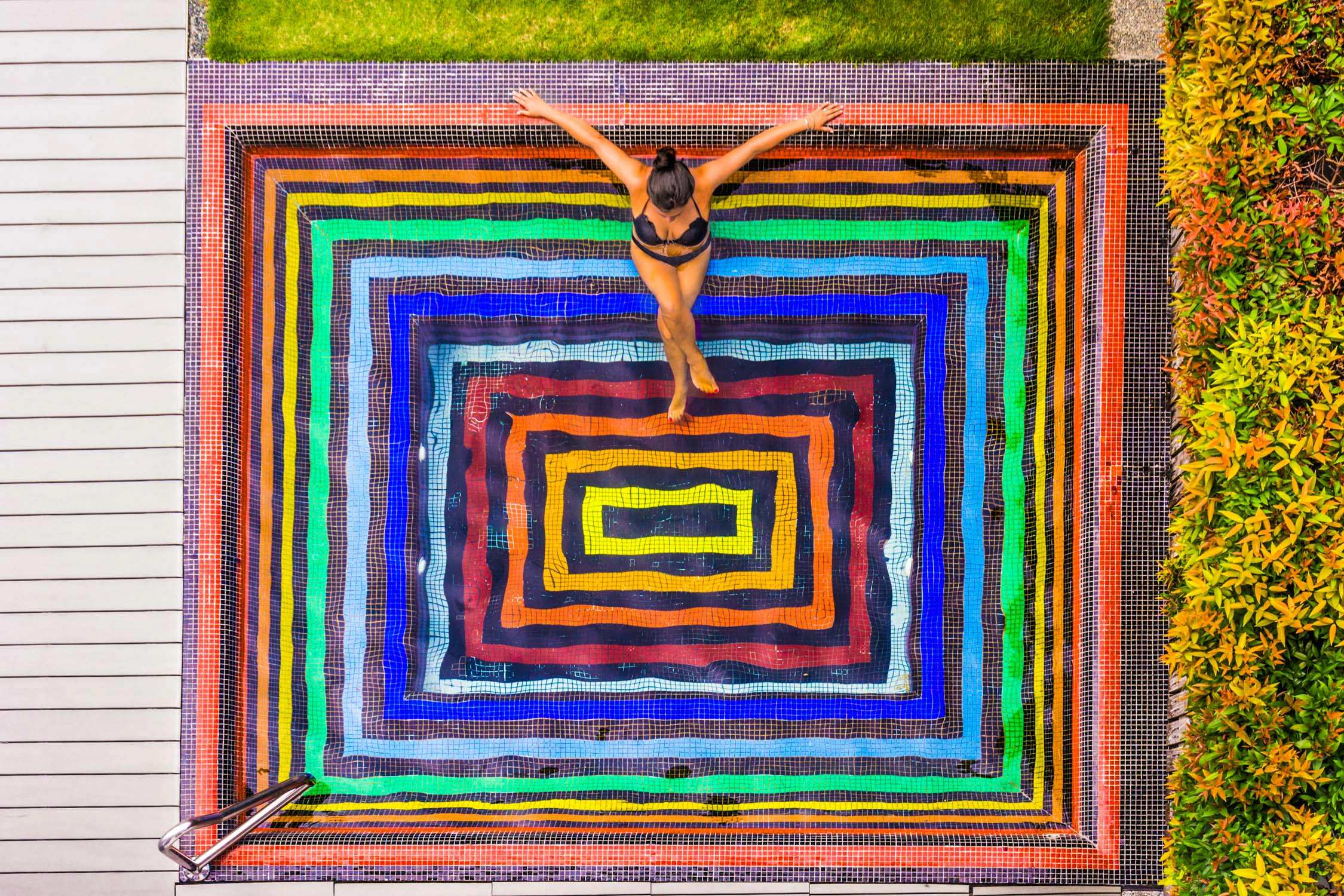 A woman lies on her back with arms outstretched over a vibrant, multi-colored rectangular pool and surrounding tiles.