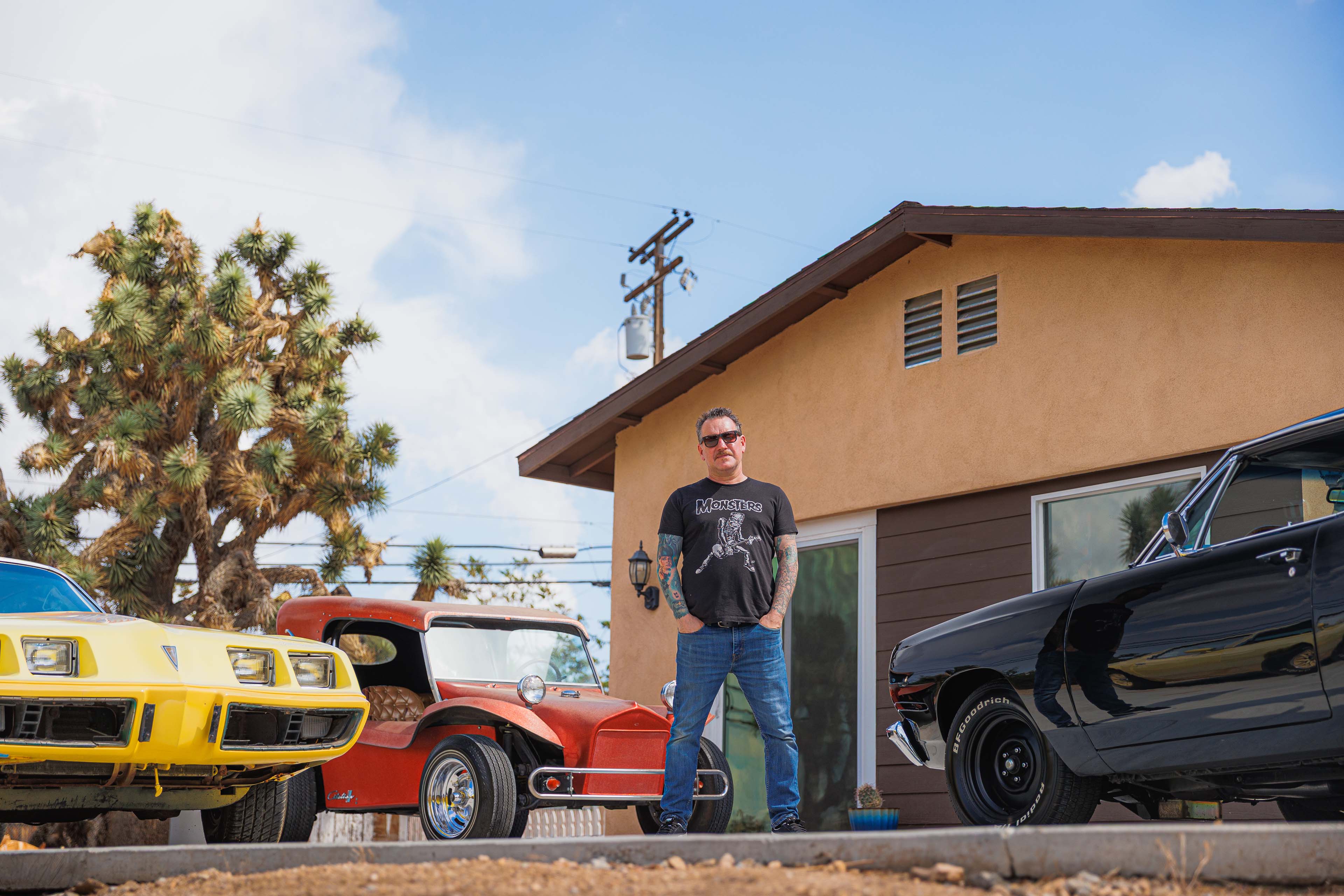 Dirty Donny Gillies standing outside his art studio wearing a “Monsters” T-shirt, flanked by a yellow Pontiac Firebird, a metallic orange Citation 2 buggy, and a black muscle car under a clear blue sky.
