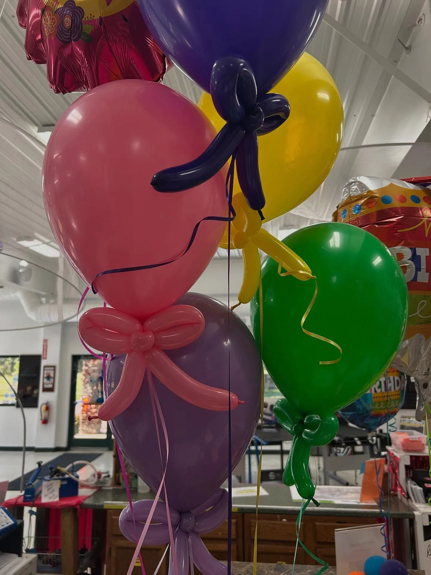Colorful assortment of helium balloons in pink, purple, green, and yellow with decorative bows, inside a festive indoor setting, ideal for celebrations and parties.
