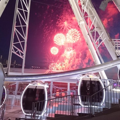 煙花在夜空中綻放，從大型摩天輪的結構內部觀賞，前景中有吊艙。