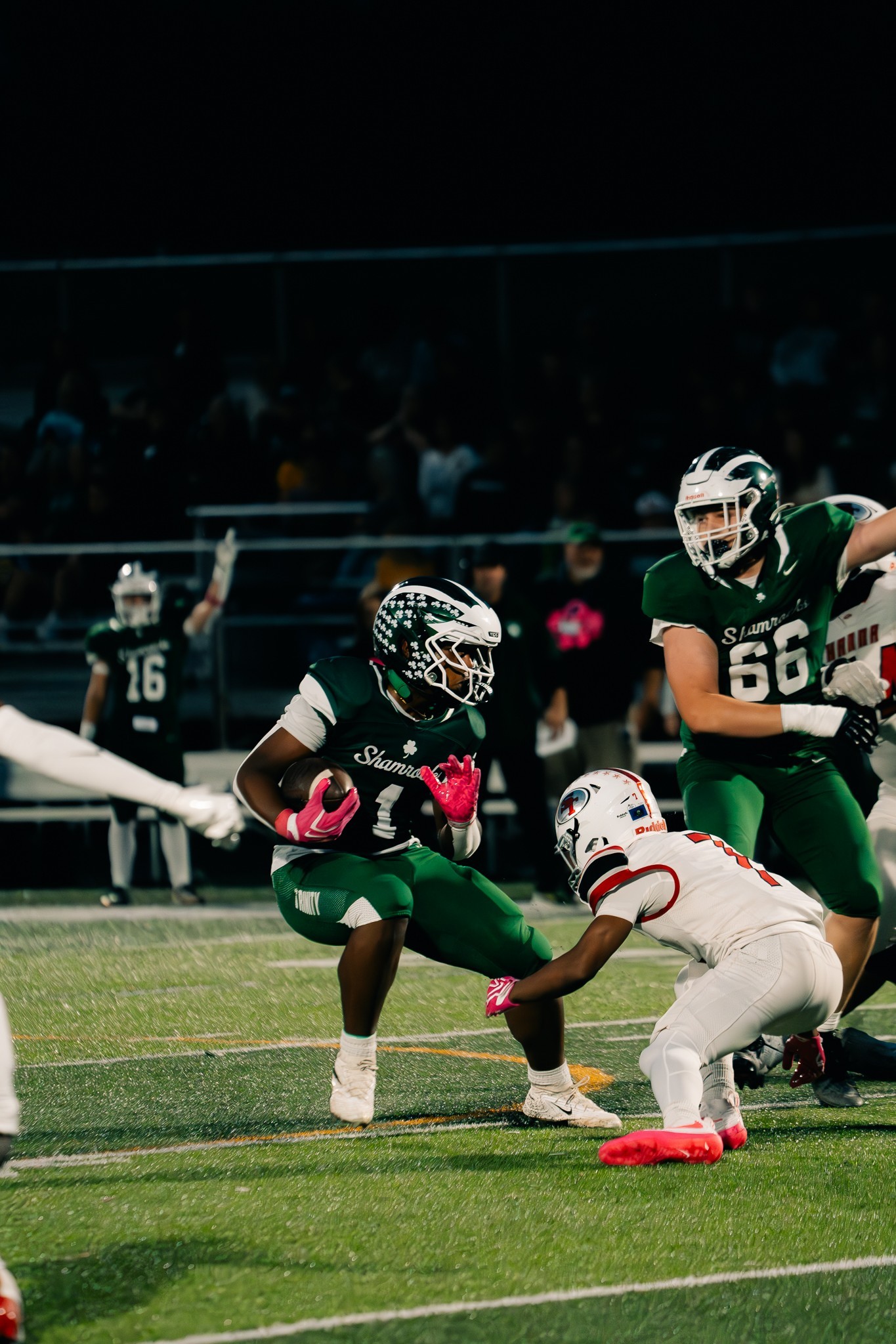 A football ball carrier in green with pink gloves faces a white-and-red defender reaching for his leg under stadium lights.