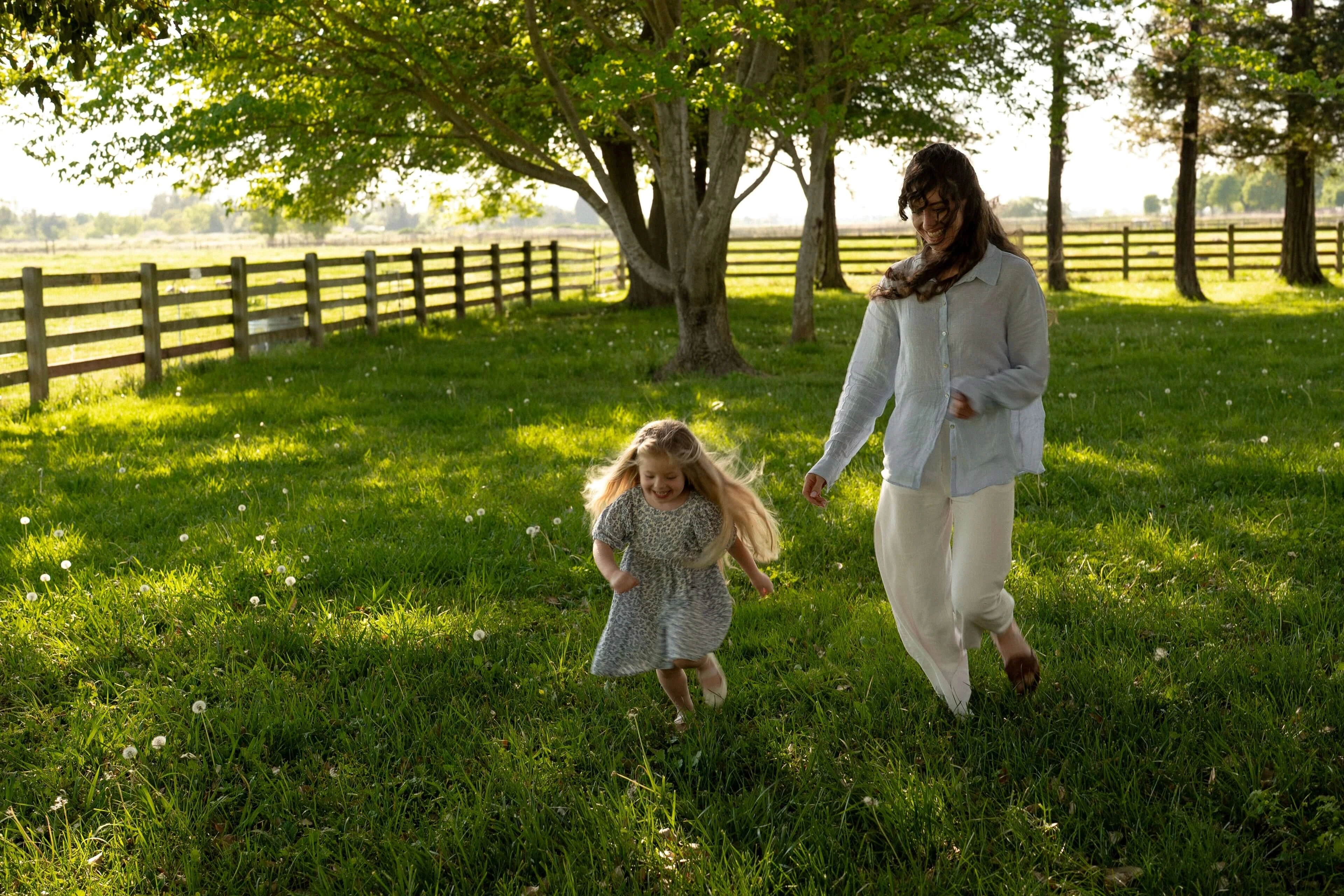 Mother and young daughter walking through a green field beneath large trees