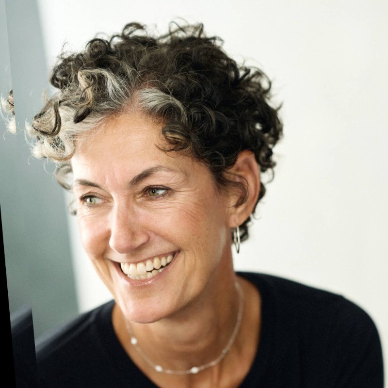 A portrait of a white woman smiling, with curly gray and black hair, wearing a silver necklace and a black t shirt