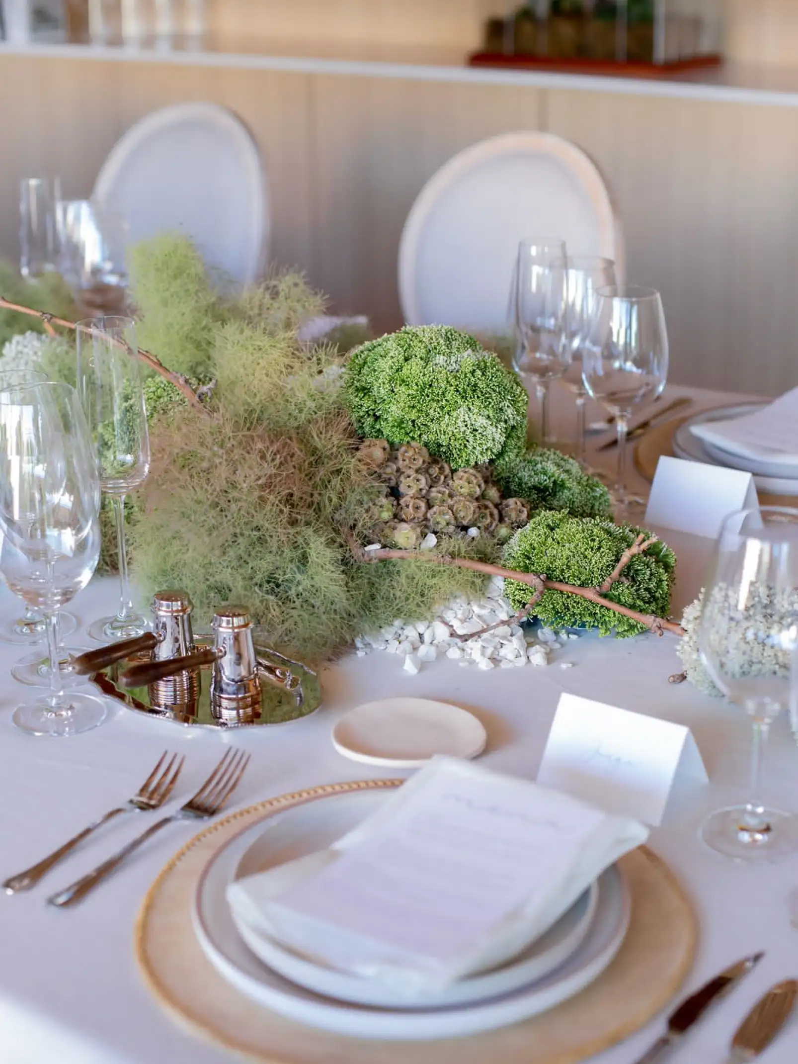 Lugar de mesa con platos blancos, servilleta y menú, rodeado de decoración vegetal minimalista en tonos verdes y neutros