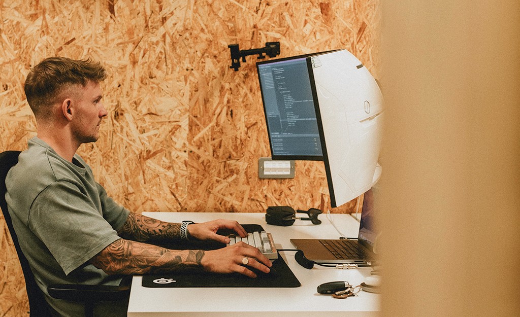 A person wearing headphones sits at a desk with a large curved monitor displaying graphic design software, surrounded by a laptop, smartphone, and potted plant, creating a modern workspace setup.