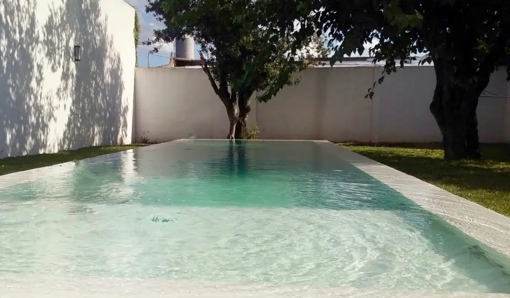 Espejo de agua cristalina en pileta de jardín revestida con material continuo Aquavations, rodeada de césped y árboles bajo la luz del sol.