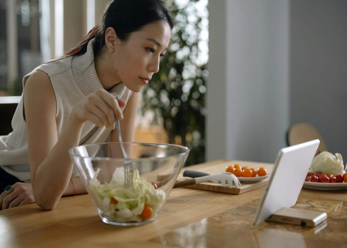 Woman eating a salad while looking at white tablet