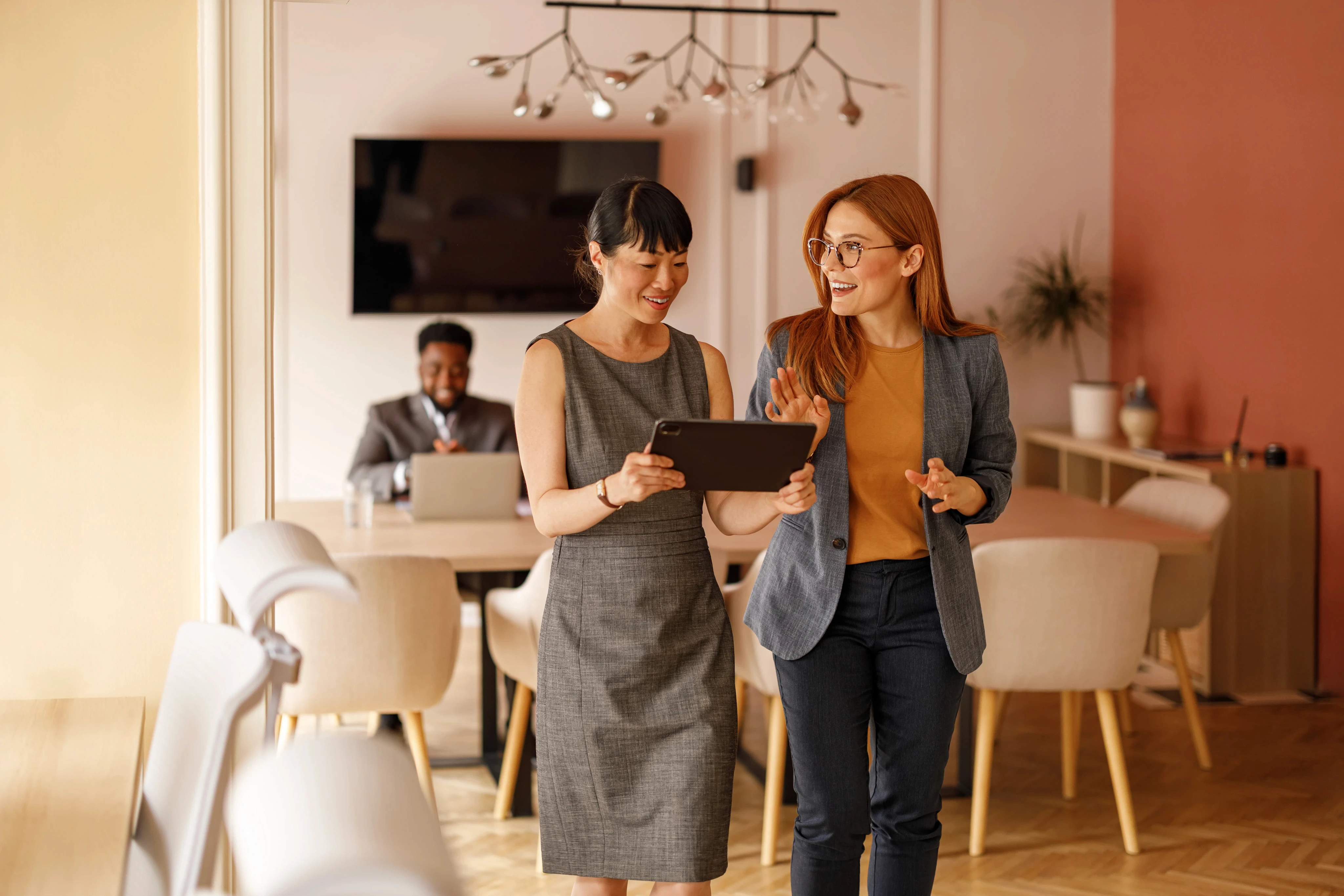Two women in business attire engage in a lively discussion, looking at a tablet, while a man works on a laptop in a modern office space.