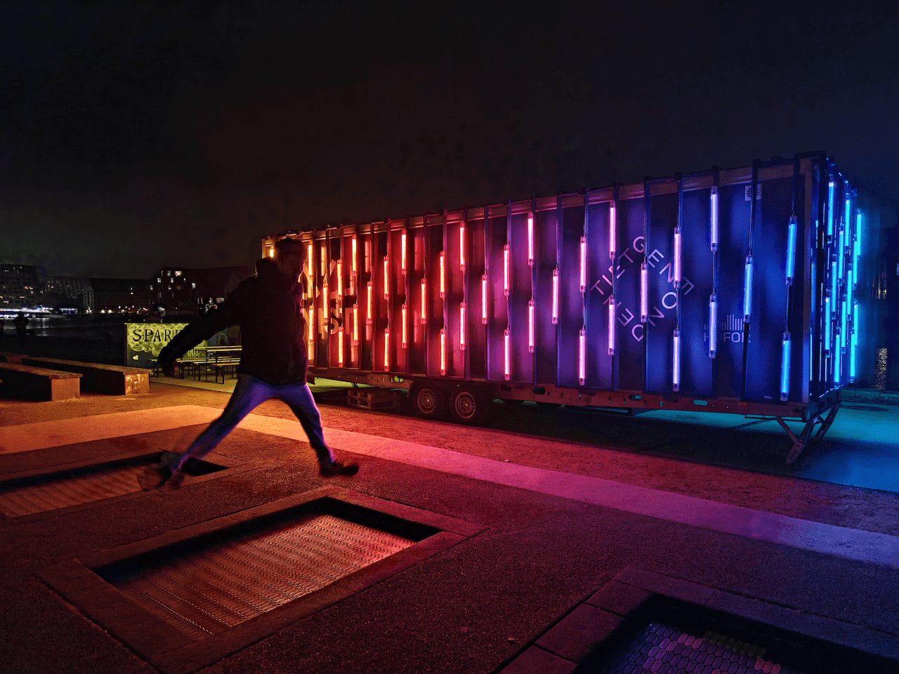 A man up in the air after jumping on a street trampoline with a colourful neon-light exhibition surrounded by darkness.