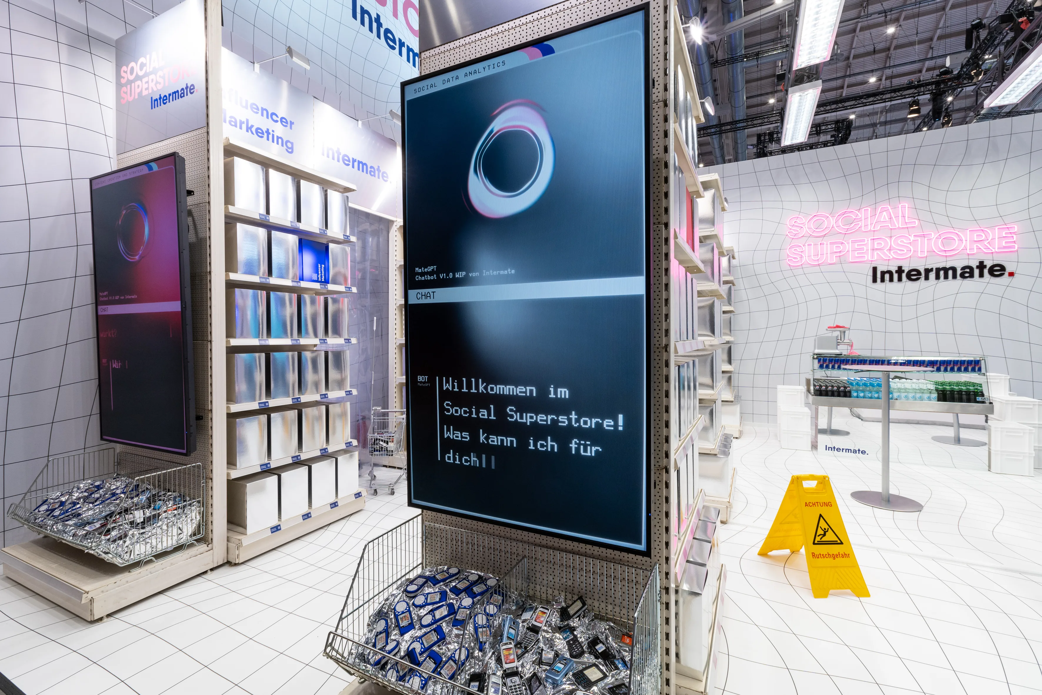 Tall chat screen welcoming visitors to the Social Superstore between shelves and baskets of old phones at the Intermate booth.
