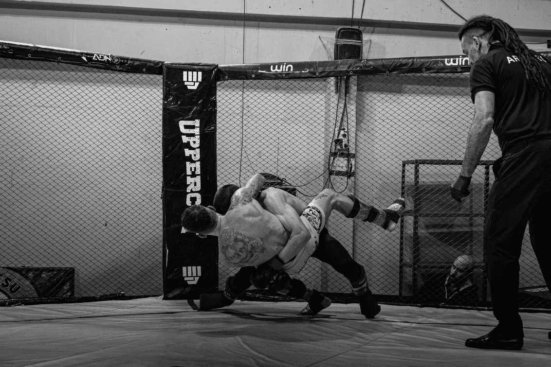 Two MMA fighters battling against the cage wall during a takedown as a referee observes.