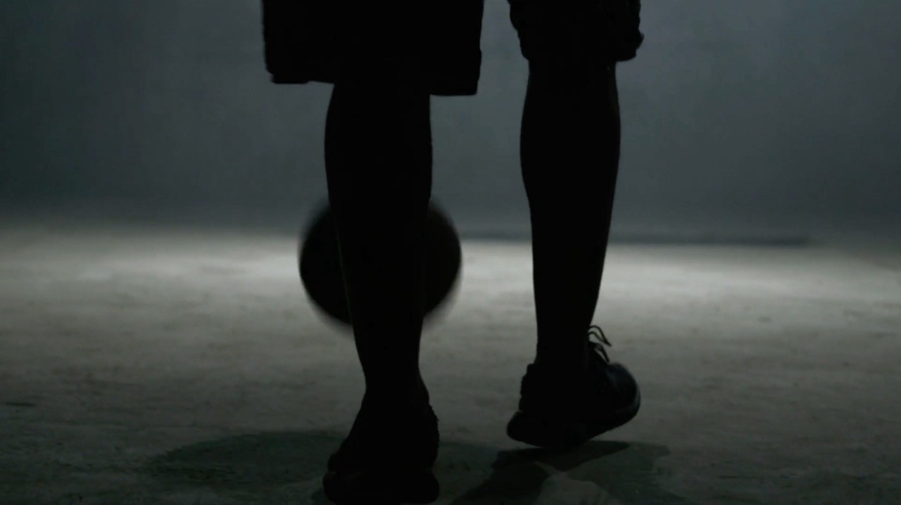 Close-up of a player's legs dribbling a basketball in a foggy, dimly lit gym.