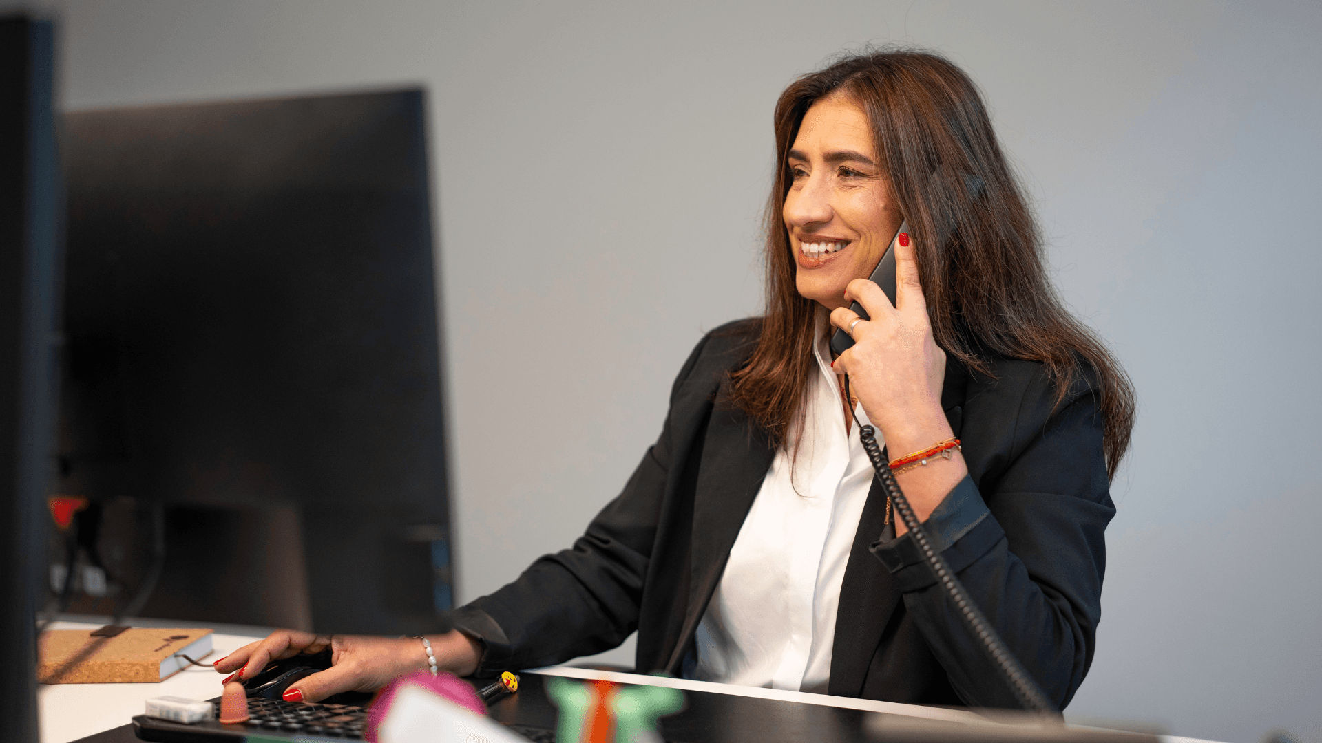 Mulher de negócios sorridente usando telefone e computador em ambiente de escritório, transmitindo profissionalismo e satisfação no trabalho.