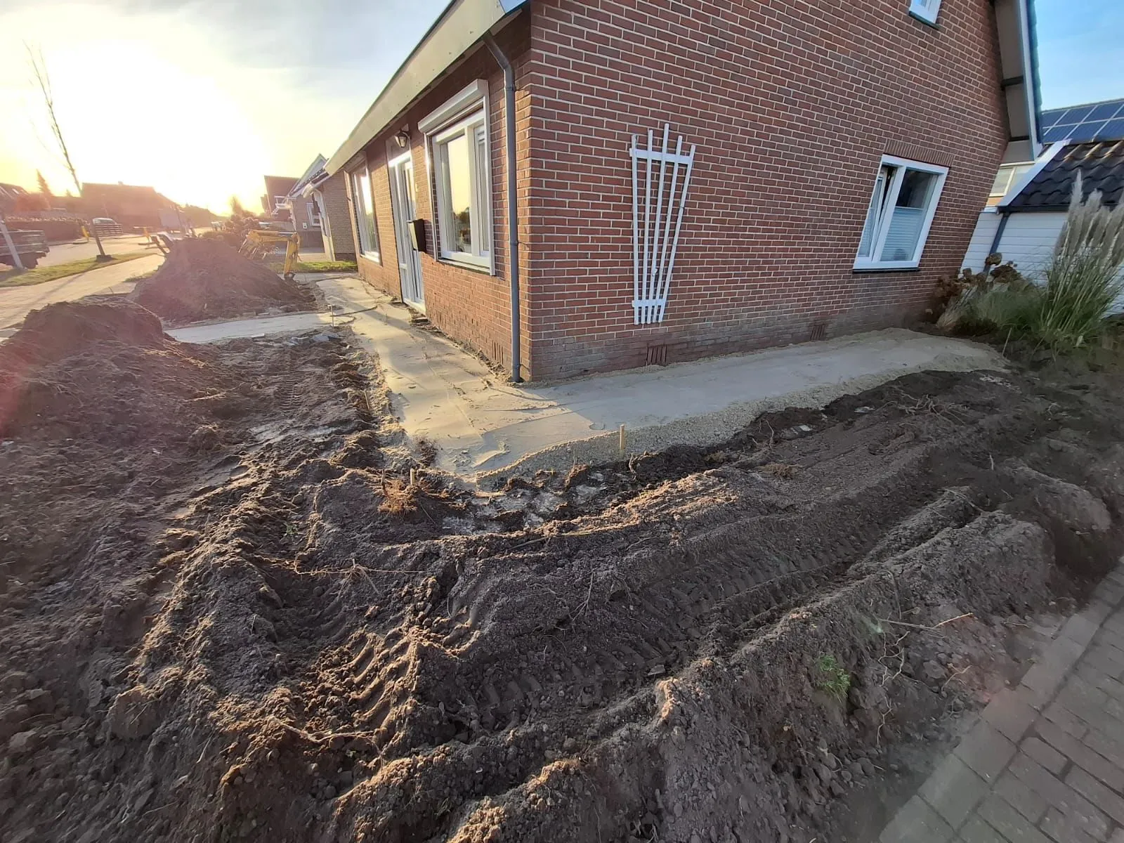 Voortuin in renovatie met nieuw bestrate paden en omgewoelde grond langs een bakstenen huis in Harkema bij ondergaande zon.
