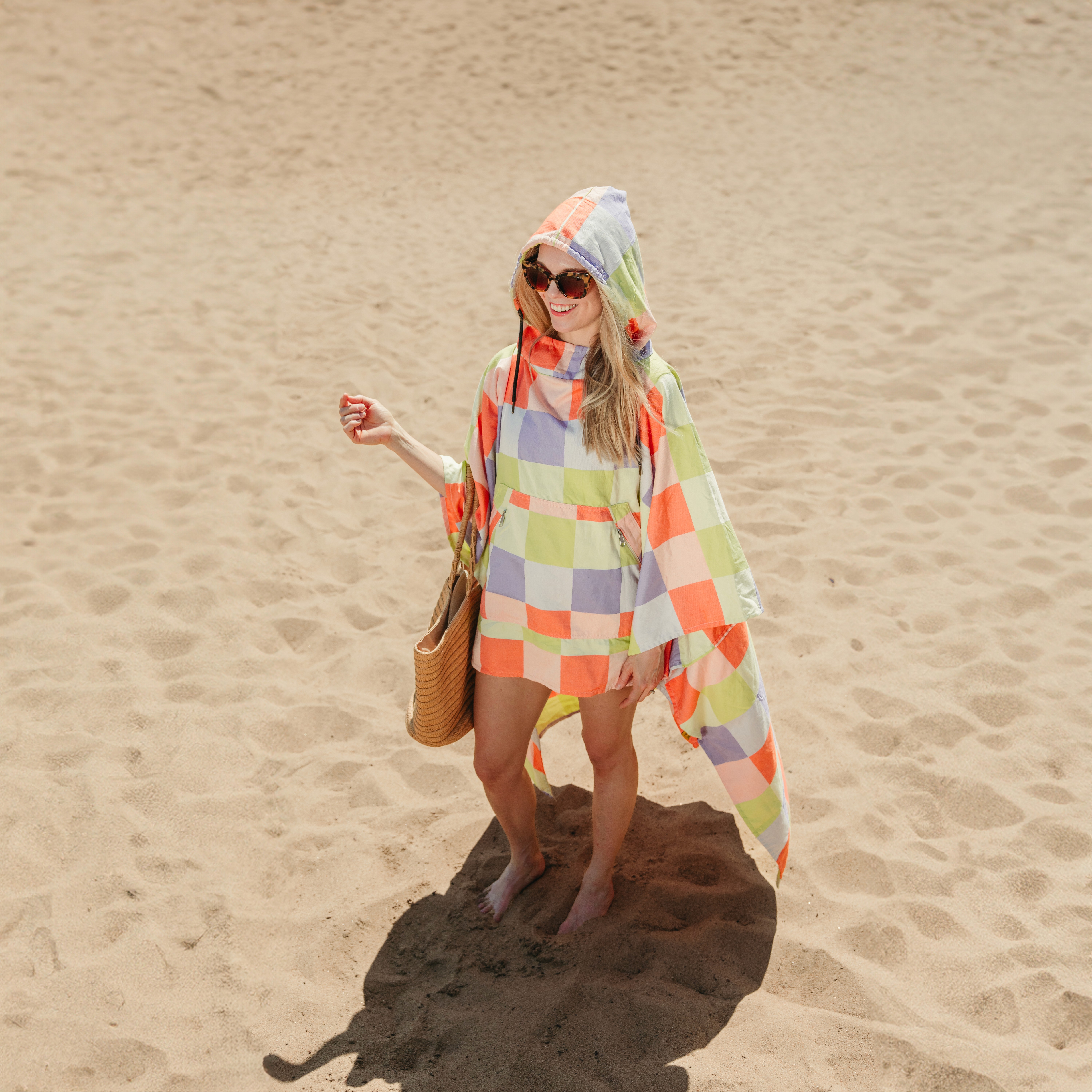 A woman walking on the sand wrapped in a pastel checkered hooded Big Blanket poncho, carrying a woven tote