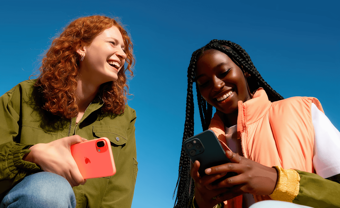 Laughing young women with red hair and braids using phones under blue sky.