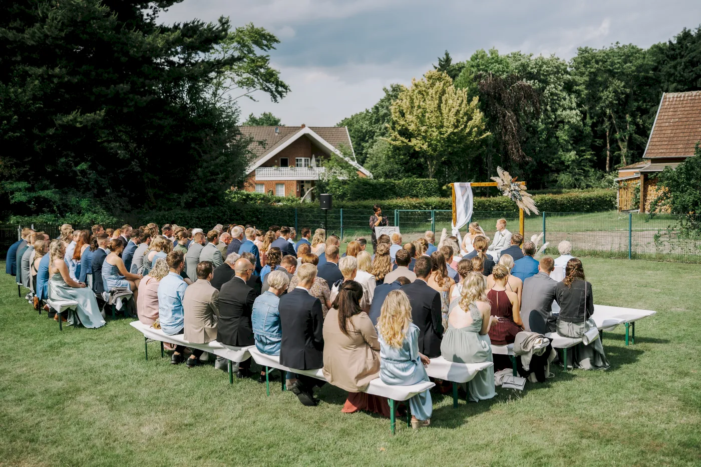 Eine Hochzeitszeremonie im Freien mit zahlreichen Gästen, die sich auf Bänken versammelt haben, um eine Zeremonie zu beobachten.