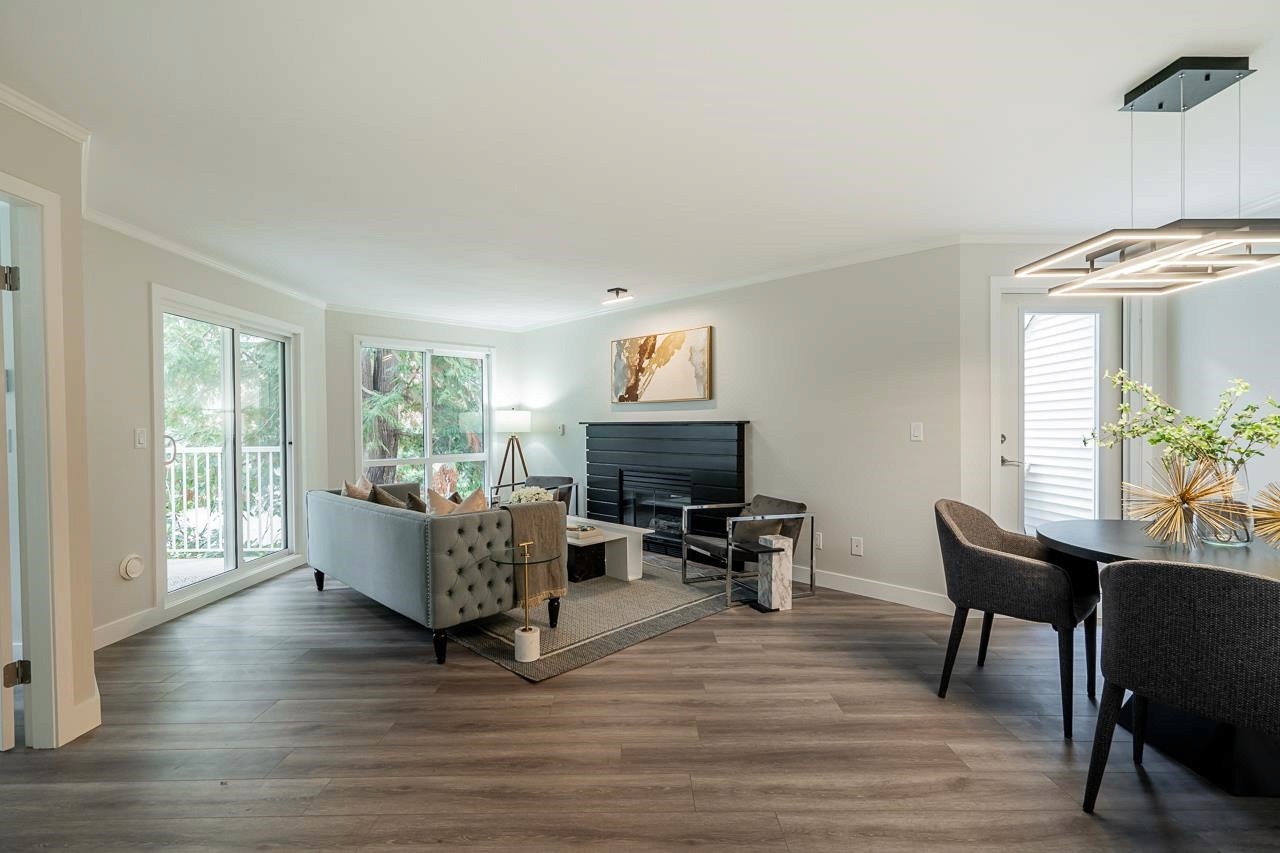 Modern living room with a gray couch, large TV, and dining area, featuring large windows and contemporary decor.