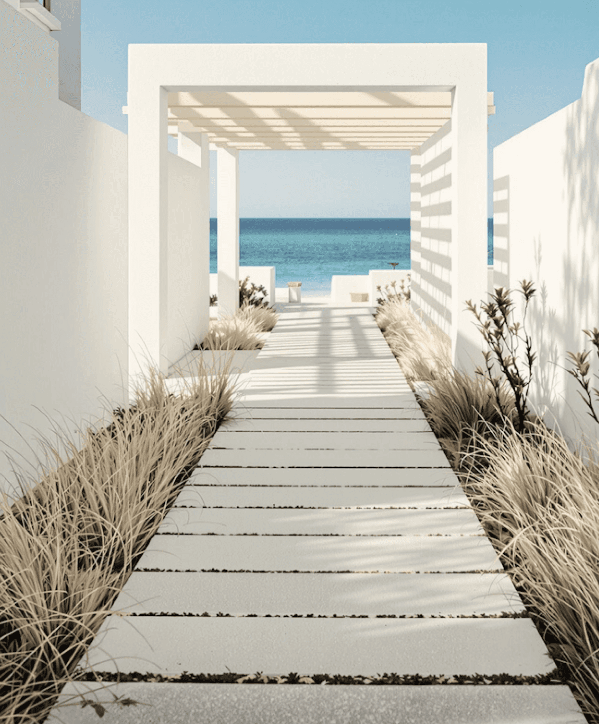 Minimalist walkway framed by white walls and pergola, leading to a serene ocean view.