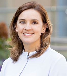 A smiling woman with shoulder-length brown hair, wearing a white coat, stands outdoors with a blurred background.