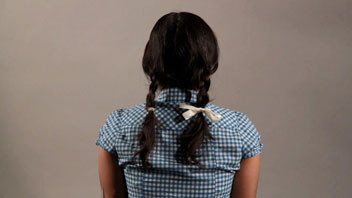 A person with dark hair styled in two braids, each secured with a small light-colored bow, is seen from behind against a plain light gray background. They are wearing a blue and white gingham short-sleeved shirt. The person’s arms are visible at the sides of their torso, and their head is turned away from the camera.