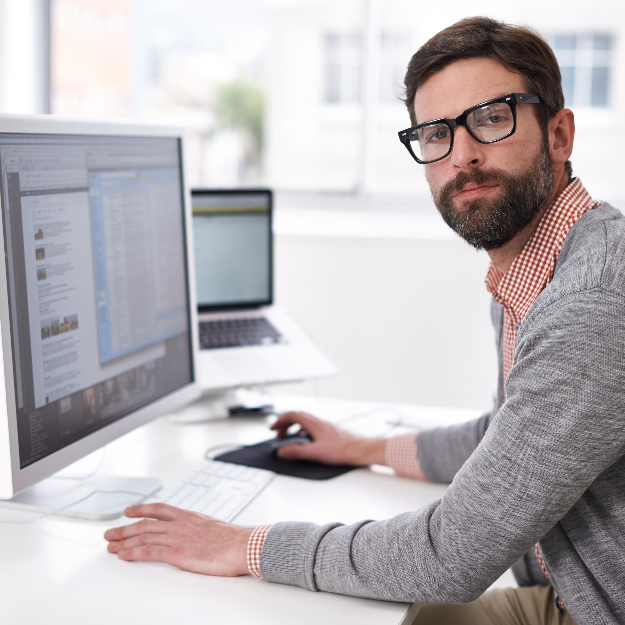 Man wearing glasses working at a desktop computer in a bright office, with a laptop open in the background.