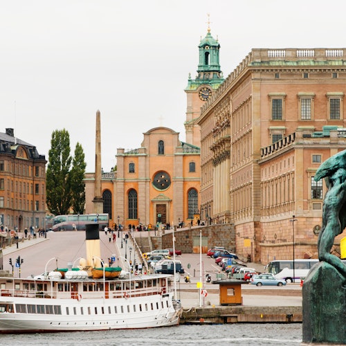 A white boat docked by a waterfront with historic buildings, including a church with a clock tower, and people walking nearby.