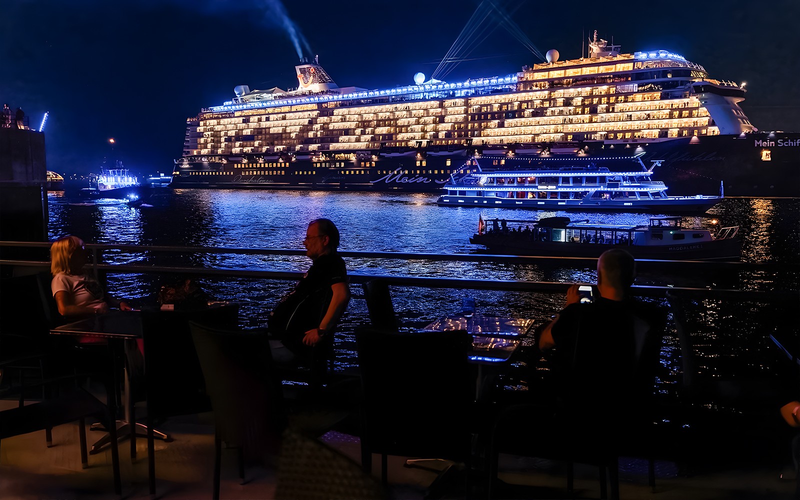 Large illuminated cruise ship on evening water, viewed from a waterfront dining area.