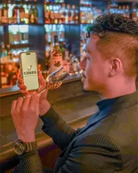 Man at a bar holding a whisky glass and phone displaying the Spiritz Explorer app among bottle-lined shelves