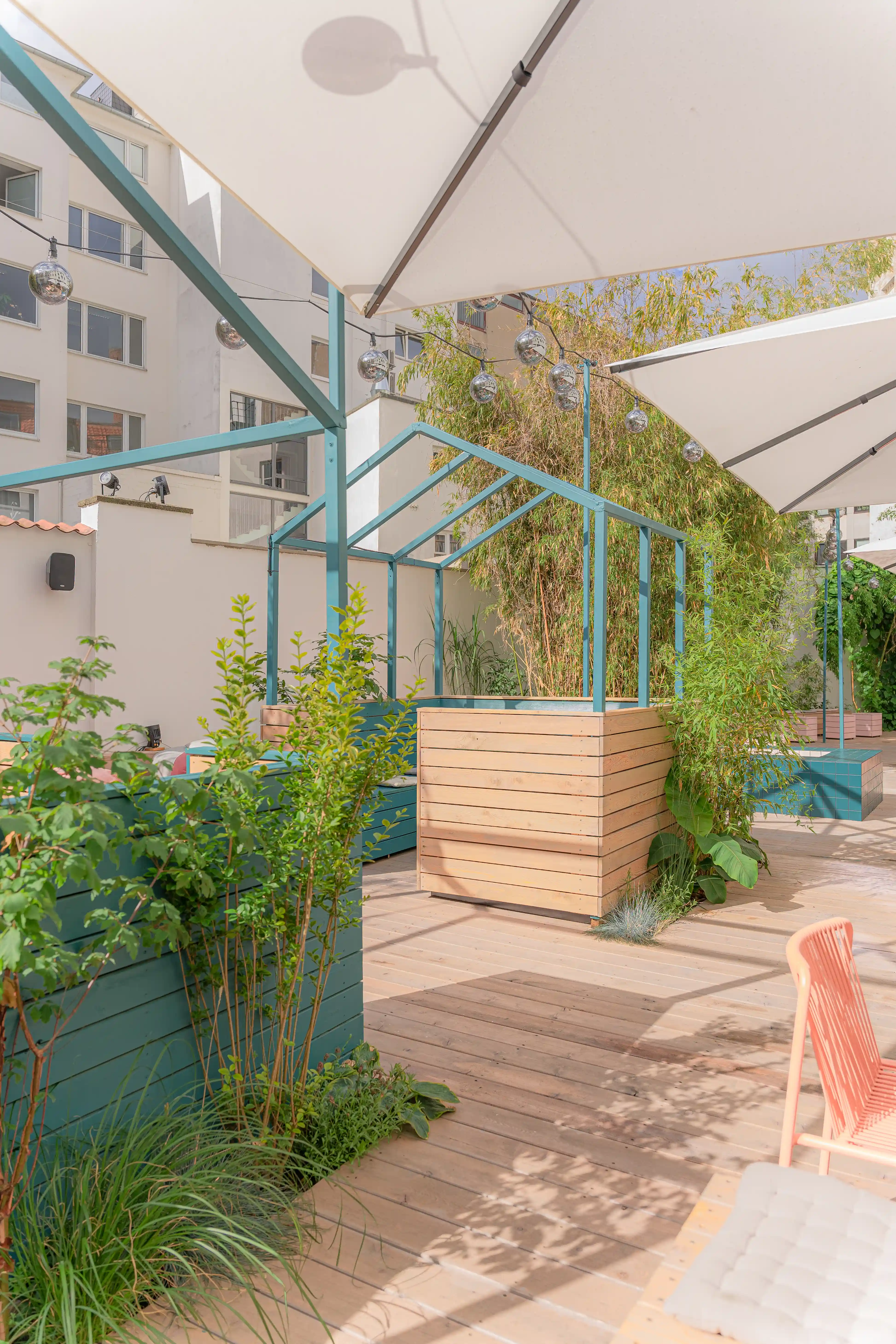 Sunny terrace at LOKAL VIER with coral-pink metal tables and chairs, wooden bench platforms, planters and large white parasols in a courtyard.