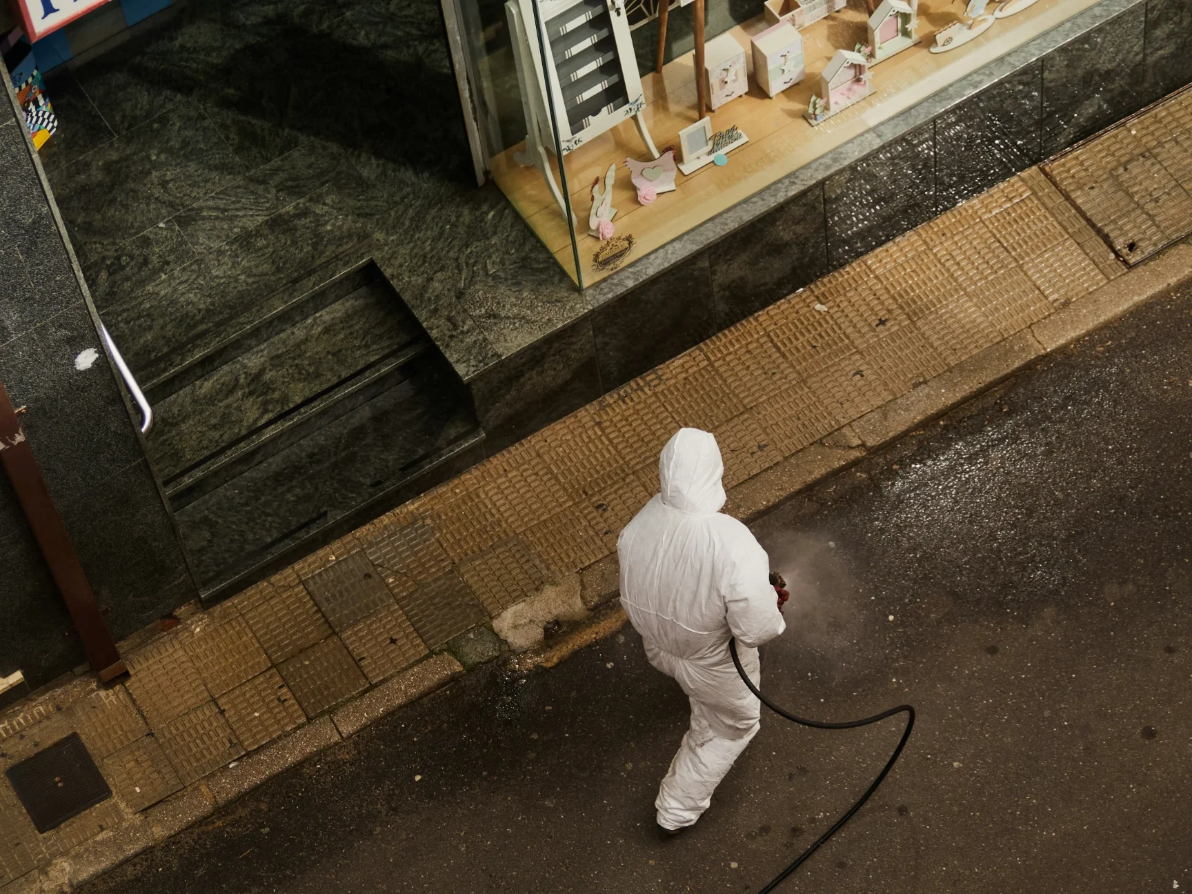 Vogelperspektive auf eine Person in einem weißen Ganzkörper-Schutzanzug, die mit einem Schlauch und einer Sprühvorrichtung den nassen Gehweg vor einem Schaufenster reinigt oder desinfiziert.