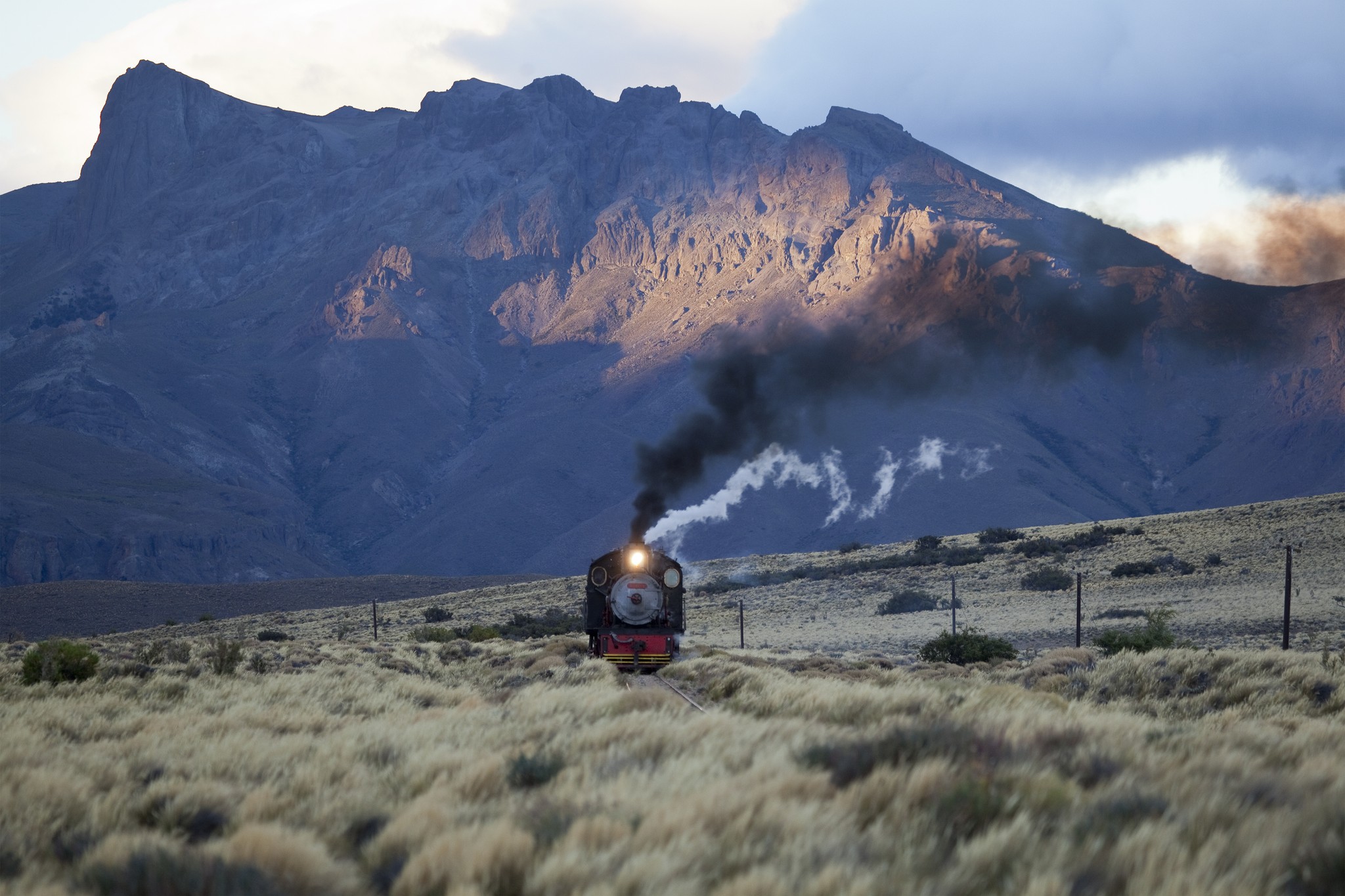 A train moves toward the viewer in the soft light of early morning, smoke billowing from its chimney, with a mountain range in the background.